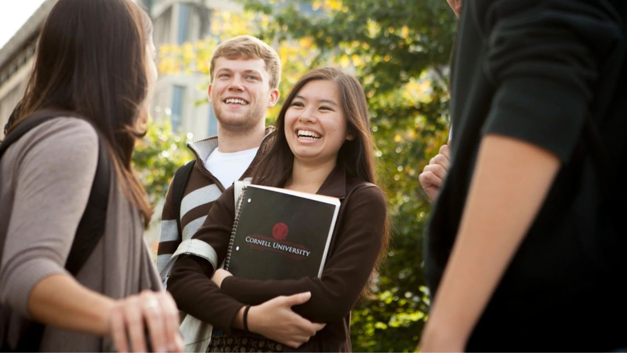 Five students stand together outside, the girl in the middle holds a Cornell University notebook.