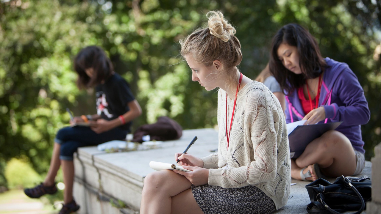Three students sit outside on a ledge, looking at forms. 