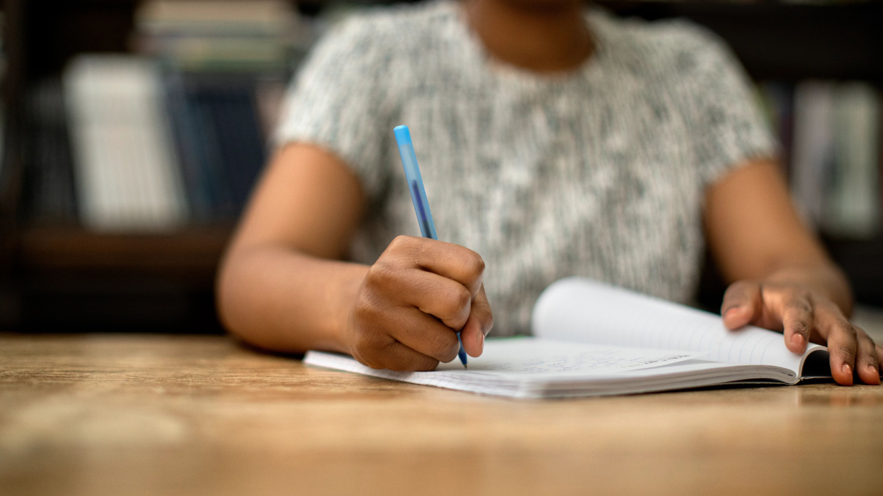 a person sits at a table, writing on a piece of paper, their body is blurred and their hand is the focus.